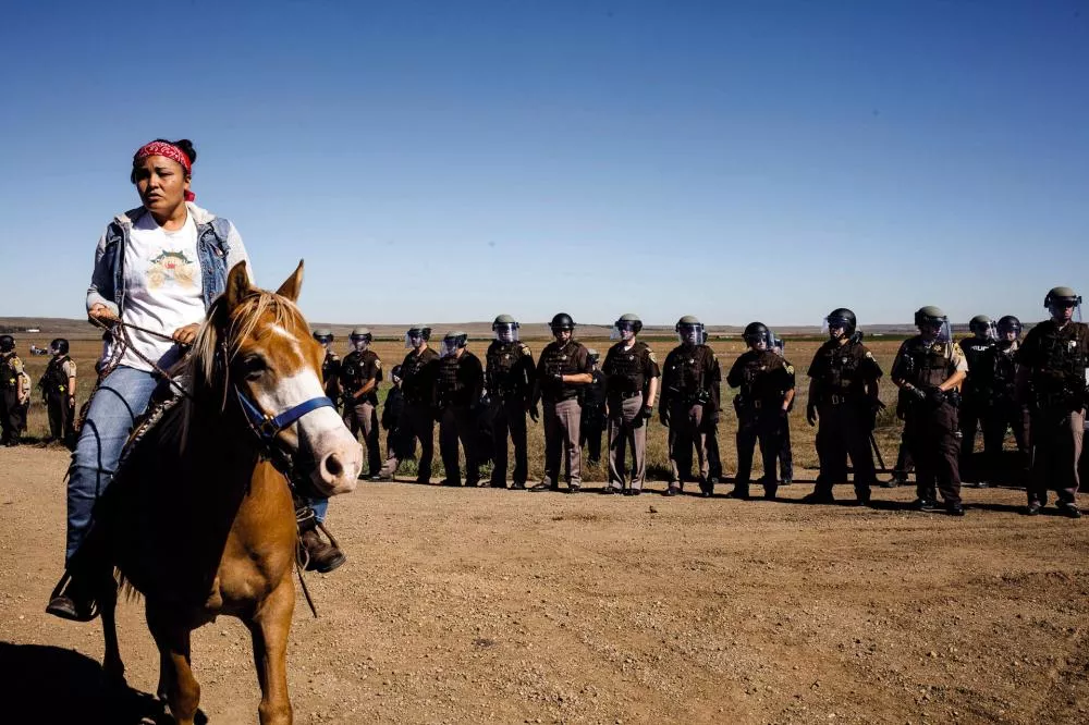 Une personne à cheval se tient devant un groupe aligné de policiers en tenue anti-émeute dans un paysage désertique.