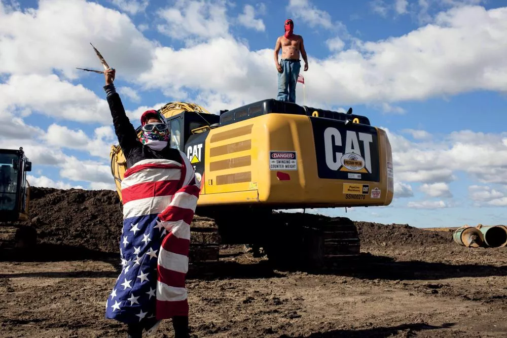 Deux personnes masquées se tiennent près d'une pelleteuse CAT sur un chantier. L'une est enveloppée dans un drapeau américain et l'autre est torse nu, debout sur la pelleteuse. Le ciel est partiellement nuageux.