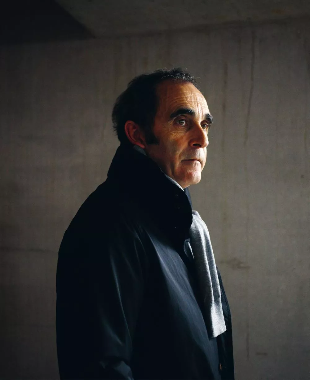 Un homme se tient debout, vêtu d'un manteau sombre et d'une écharpe grise, regardant vers la droite. L'arrière-plan est sobre et minimaliste, avec un mur de couleur neutre.