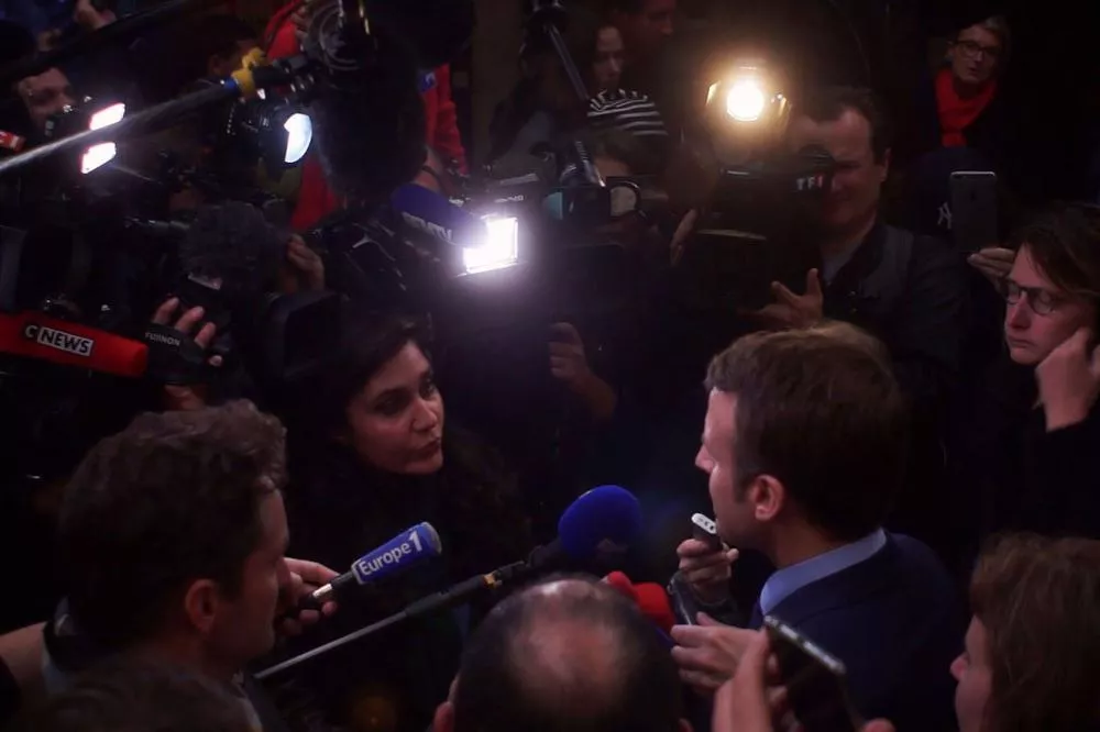 Un homme entouré de nombreux journalistes et caméras, répondant à leurs questions lors d'une conférence de presse ou d'un événement médiatique.