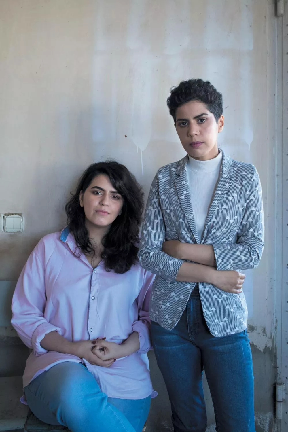 Deux personnes sont debout devant un mur clair. L'une porte une chemise rose et est assise, tandis que l'autre, debout, porte un blazer à motifs avec un col roulé blanc. Elles ont toutes les deux les bras croisés ou posés.