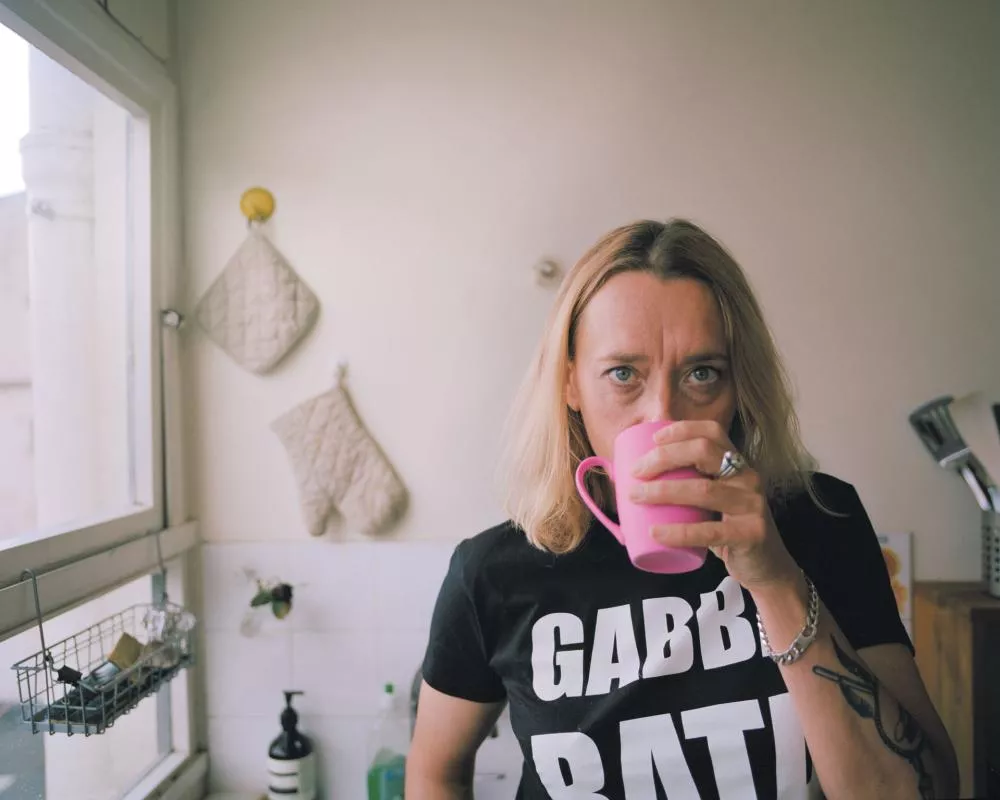 Une personne blonde se tient dans une cuisine, tenant une tasse rose. Elle porte un t-shirt noir avec des lettres blanches. Des gants de cuisine sont accrochés au mur derrière elle.