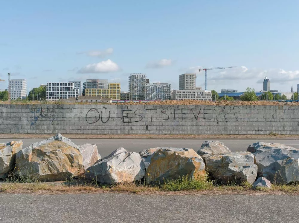 Un mur en béton avec un graffiti disant "OÙ EST STEVE???" en grandes lettres noires. Des bâtiments modernes et une grue sont visibles en arrière-plan. Des rochers se trouvent au premier plan.