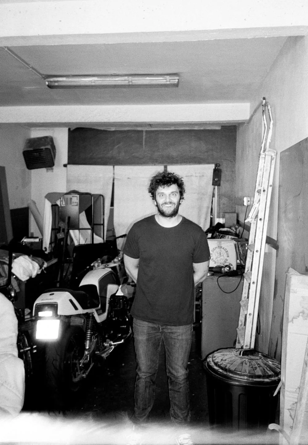 Un homme se tient debout dans un garage ou un atelier, avec des motos et divers objets en arrière-plan.