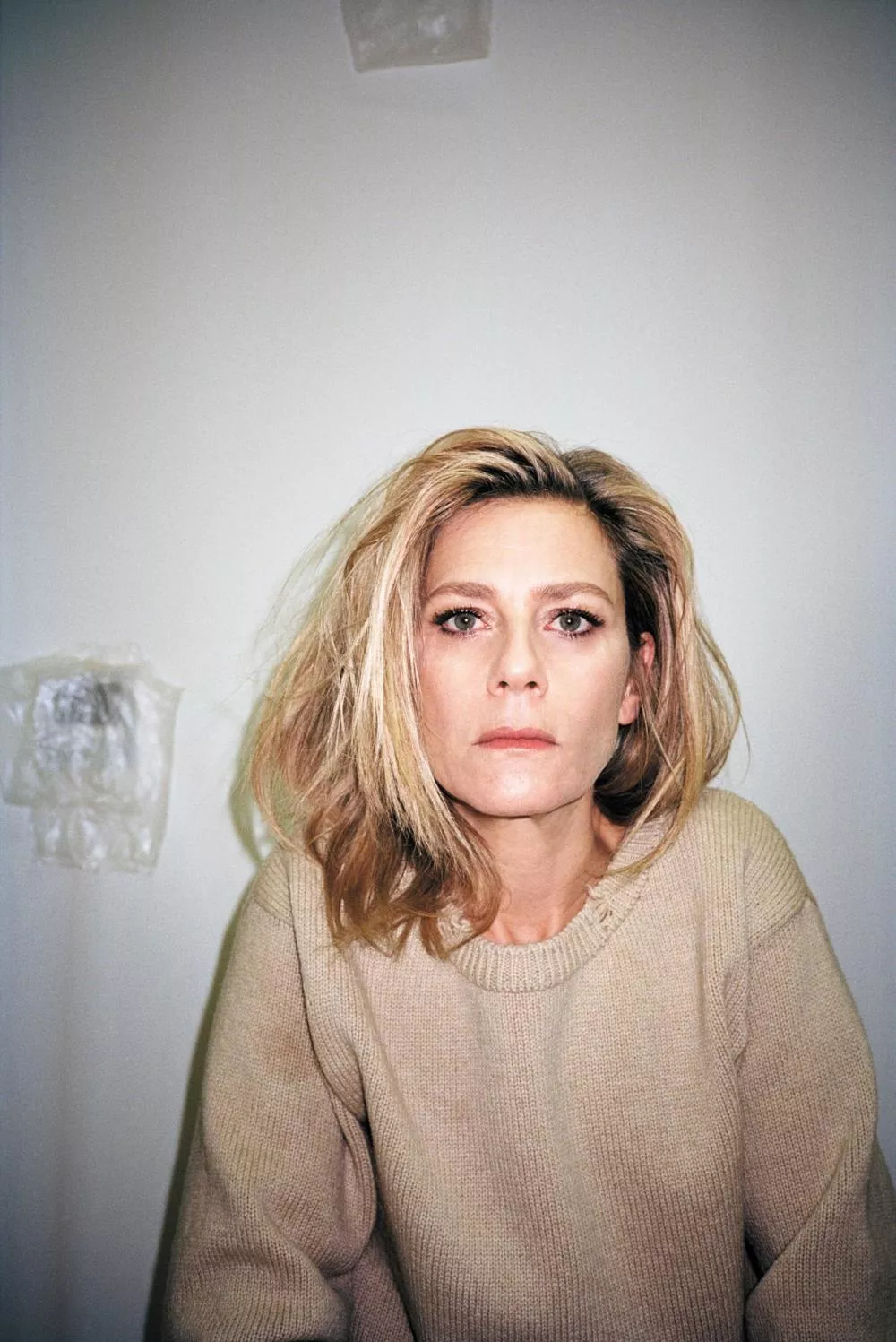 Une personne aux cheveux blonds et au regard intense porte un pull beige. L'arrière-plan est neutre avec des éléments flous.