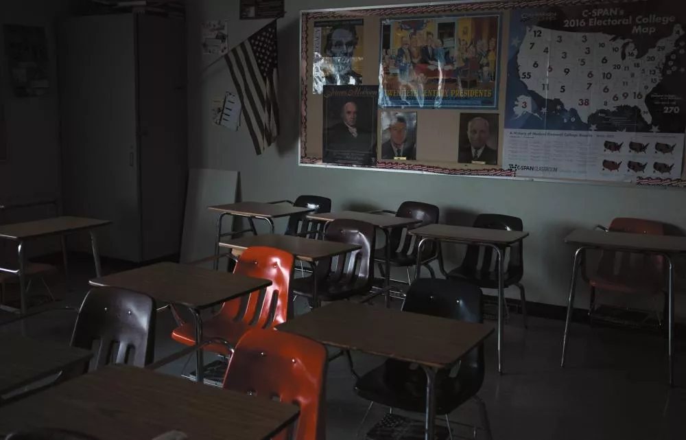 Une salle de classe vide avec des bureaux et des chaises, un drapeau américain accroché au mur, et des affiches éducatives, y compris une carte électorale des États-Unis de 2016.