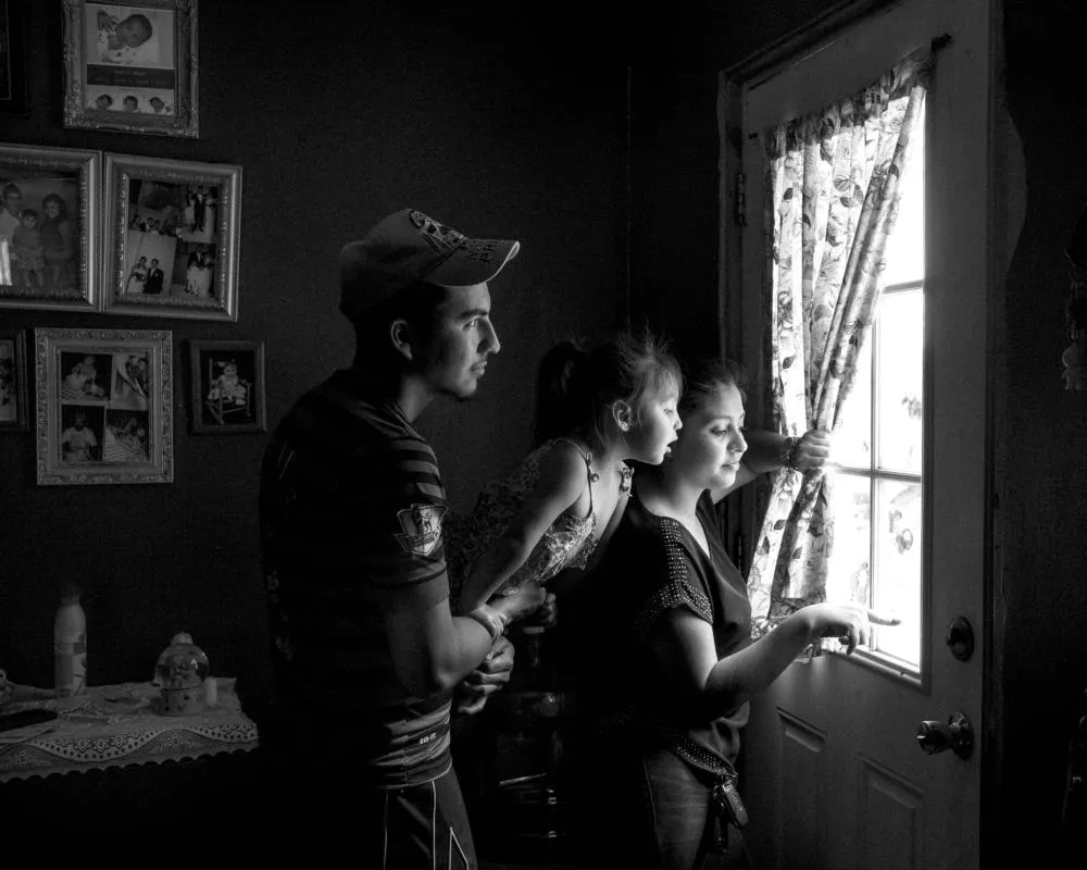 Une famille composée de trois personnes, dont un homme, une femme et une petite fille, regarde par une porte vitrée. Ils semblent attentifs à ce qui se passe à l'extérieur. Des cadres photo sont accrochés au mur à gauche. L'image est en noir et blanc.