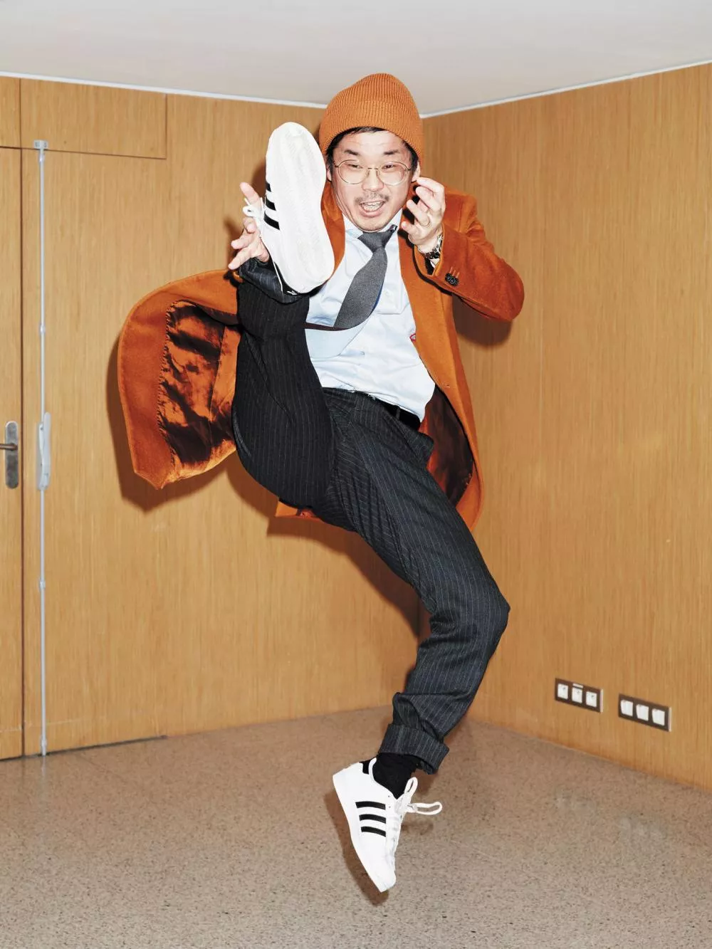 Un homme portant un manteau orange, un bonnet assorti, une chemise bleue, une cravate grise et des baskets blanches avec des bandes noires effectue un saut dynamique dans une pièce aux murs en bois.