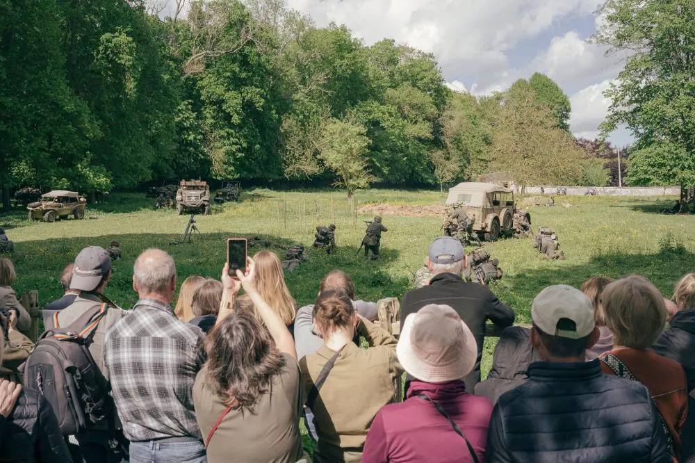 Un groupe de personnes observe une reconstitution historique militaire en plein air, avec des véhicules anciens et des participants en tenue militaire.