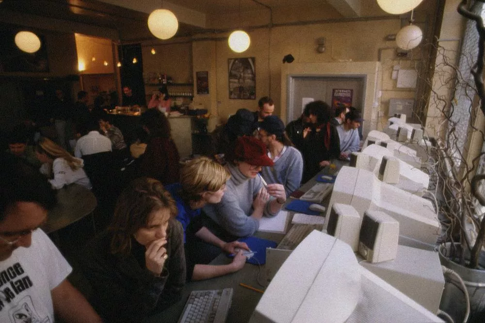 Un groupe de personnes est assis devant une rangée d'ordinateurs dans un café internet. Certaines personnes discutent, tandis que d'autres semblent concentrées sur leurs écrans. L'ambiance est animée et conviviale.
