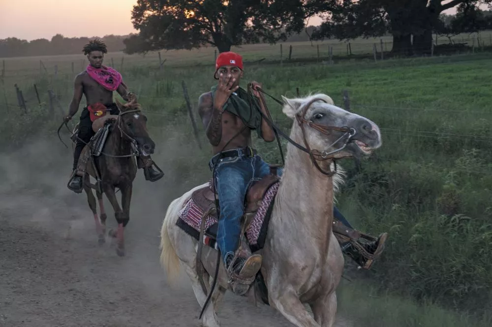 Deux cavaliers montent des chevaux sur un chemin de terre. Le cavalier de droite porte une casquette rouge, un jean et est torse nu, tenant les rênes d'une main et faisant un signe avec l'autre. Le cavalier à gauche est également torse nu, avec un foulard rose autour du cou, et tient les rênes de son cheval. L'arrière-plan montre un paysage de campagne avec des arbres et des clôtures.