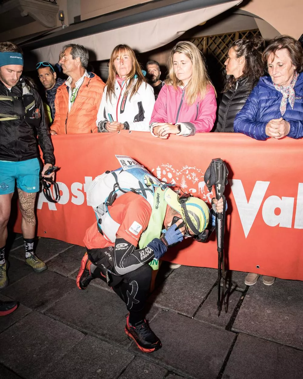 Un coureur s'agenouille, appuyé sur des bâtons de marche, devant une barrière où se tiennent plusieurs spectateurs.