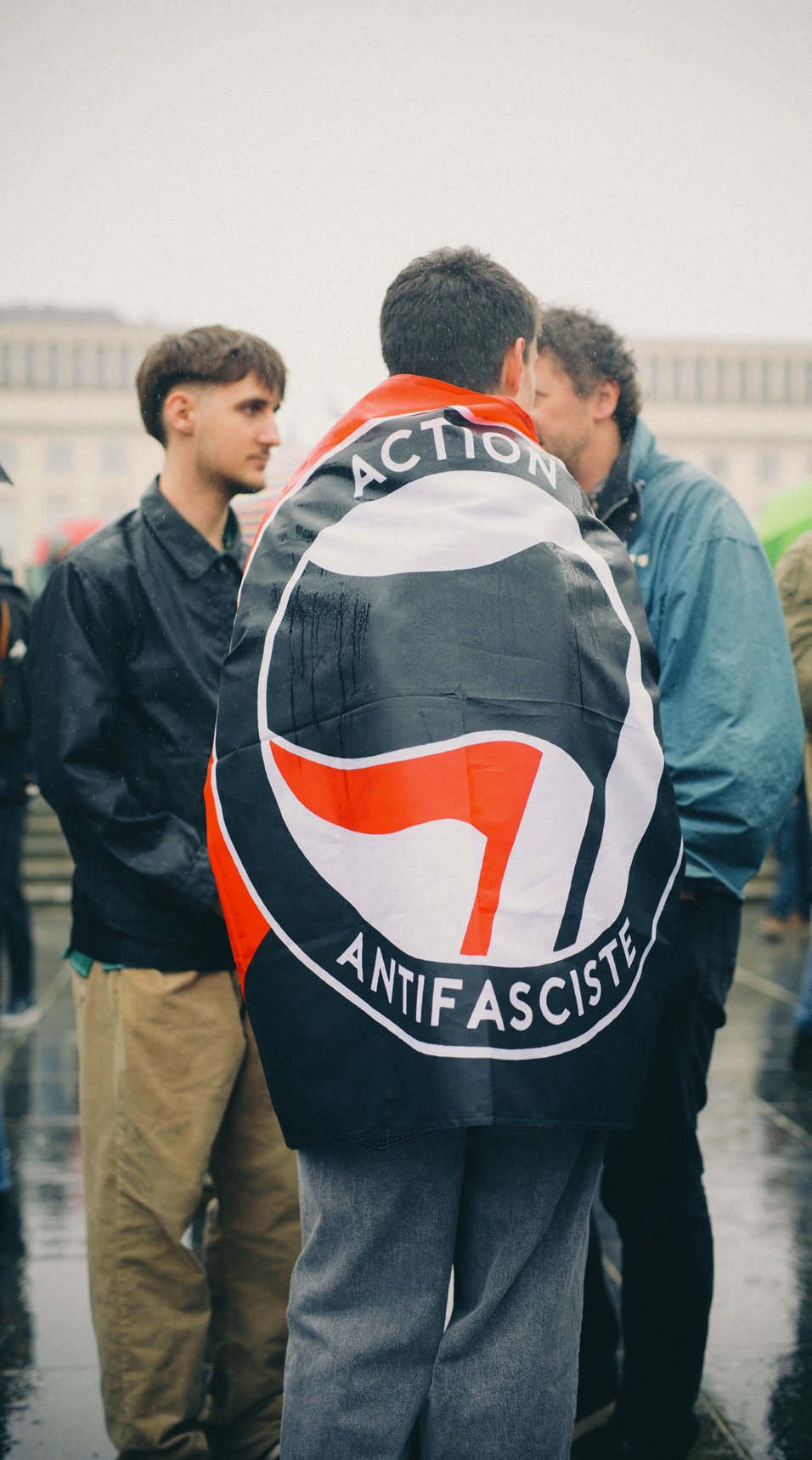 Un individu porte un drapeau avec le symbole et le texte "Action Antifasciste" lors d'une manifestation ou d'un rassemblement.