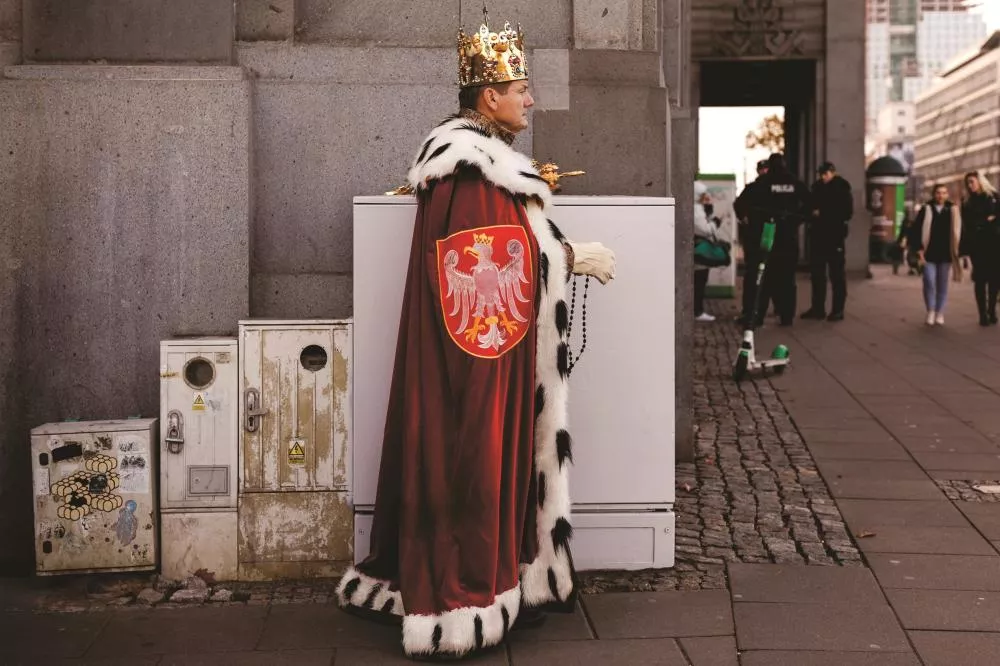 Un homme portant une couronne et une cape royale se tient debout dans une rue, adossé à une structure en béton. La cape est ornée d'un blason avec un aigle. En arrière-plan, des passants et des policiers sont visibles.