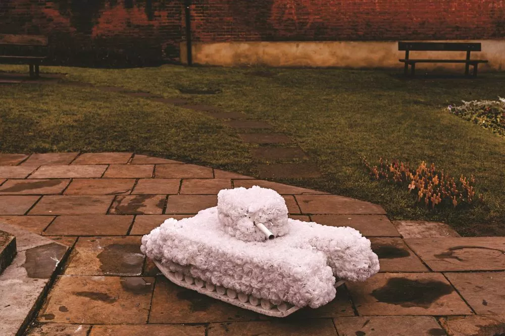 Un char miniature recouvert de fleurs blanches est posé sur un sol pavé dans un jardin, avec des bancs et des petites croix en arrière-plan.