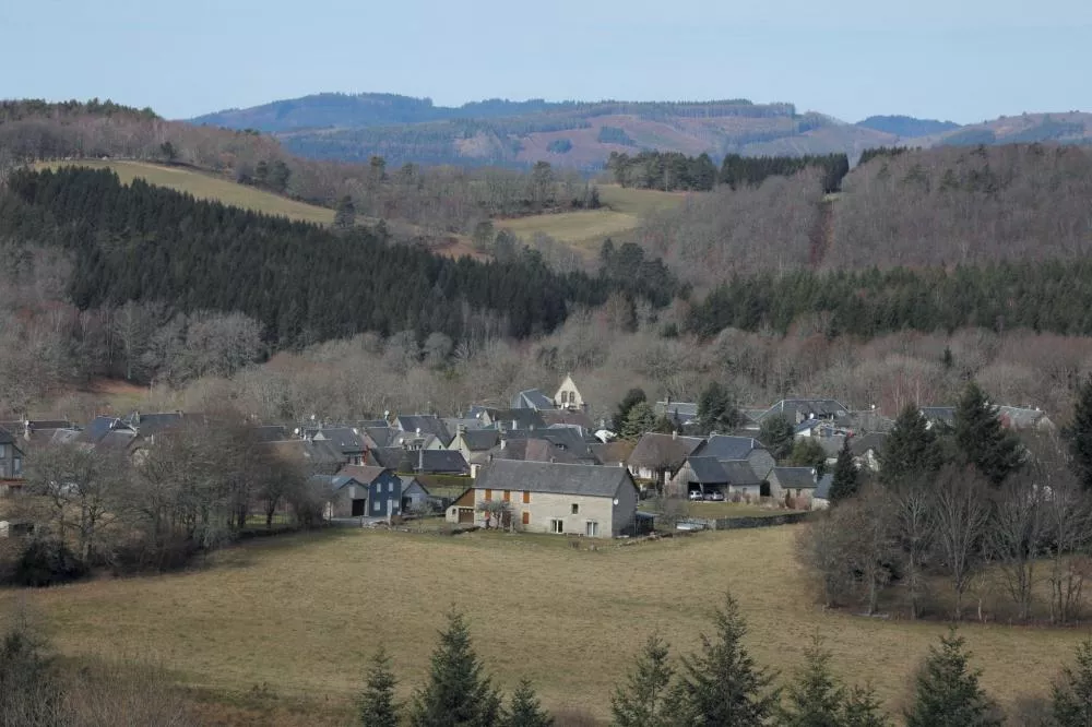 Un village rural entouré de collines boisées et de prairies, avec des maisons en pierre et un paysage naturel en arrière-plan.
