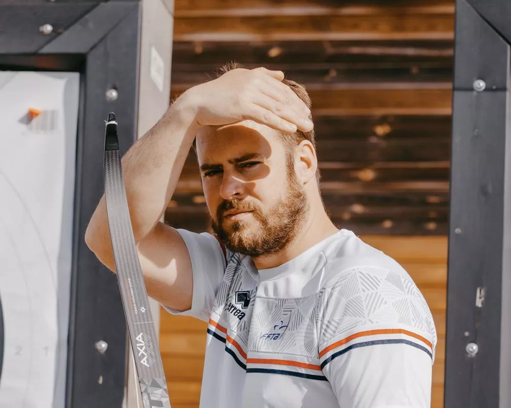 Un homme avec une barbe, portant un t-shirt blanc avec des rayures, se tient près d'une structure en bois et se protège les yeux du soleil avec sa main.