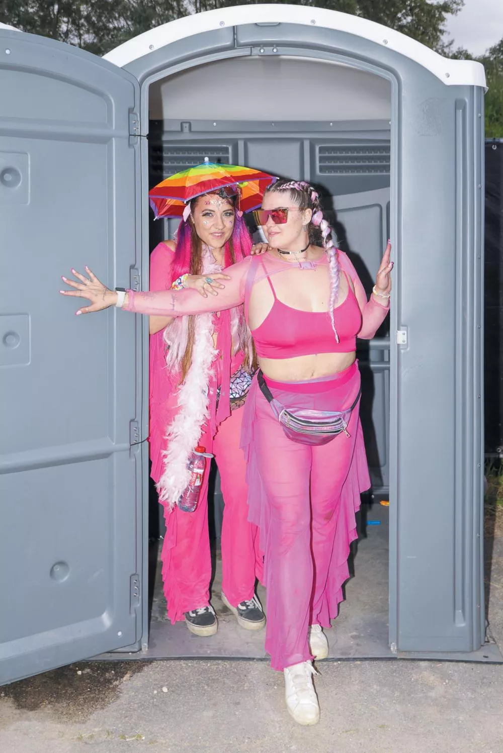 Deux personnes portant des tenues roses sortent d'une cabine de toilettes portative. L'une d'elles porte un parapluie arc-en-ciel et des accessoires colorés.