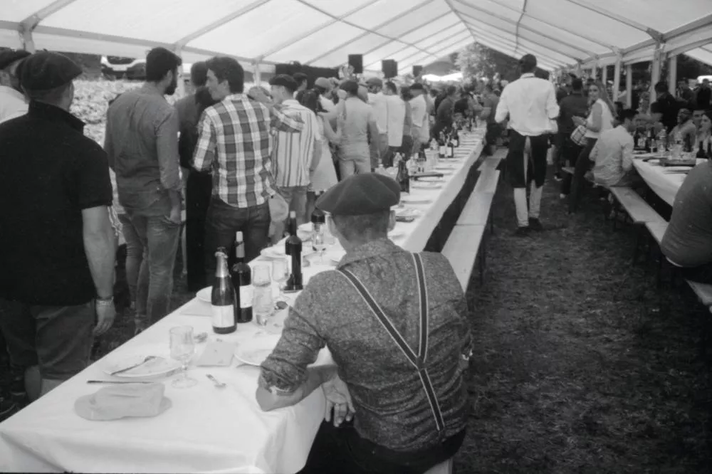Un grand repas en plein air sous une tente, avec des tables alignées et des personnes assises ou debout, certaines portant des bérets. Les tables sont couvertes de nappes blanches et de bouteilles de vin.