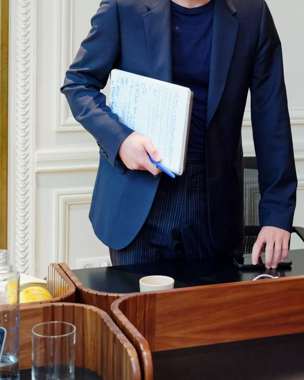 Un homme en costume bleu marine tient un carnet avec des notes manuscrites et un stylo bleu. Il se tient près d'une table avec des objets comme une bouteille d'eau, une tasse, et un téléphone.