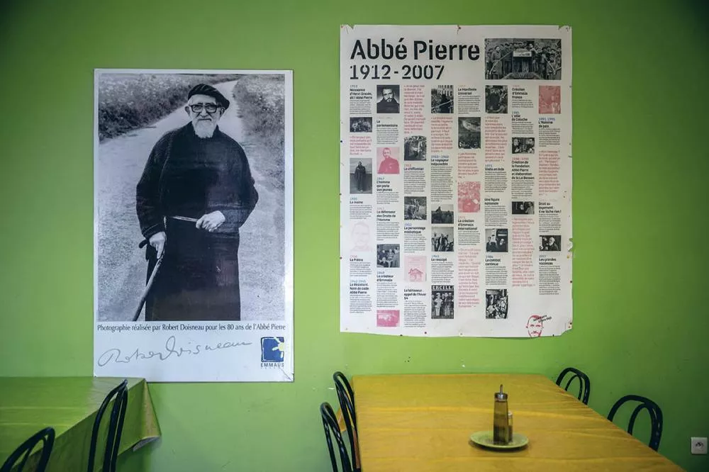 Un mur vert avec deux affiches : à gauche, une photo en noir et blanc d'un homme âgé avec une canne, signée par Robert Doisneau pour les 80 ans de l'Abbé Pierre ; à droite, une affiche détaillant la biographie de l'Abbé Pierre de 1912 à 2007. Au premier plan, une table jaune avec des chaises noires et une bouteille d'huile sur une assiette.