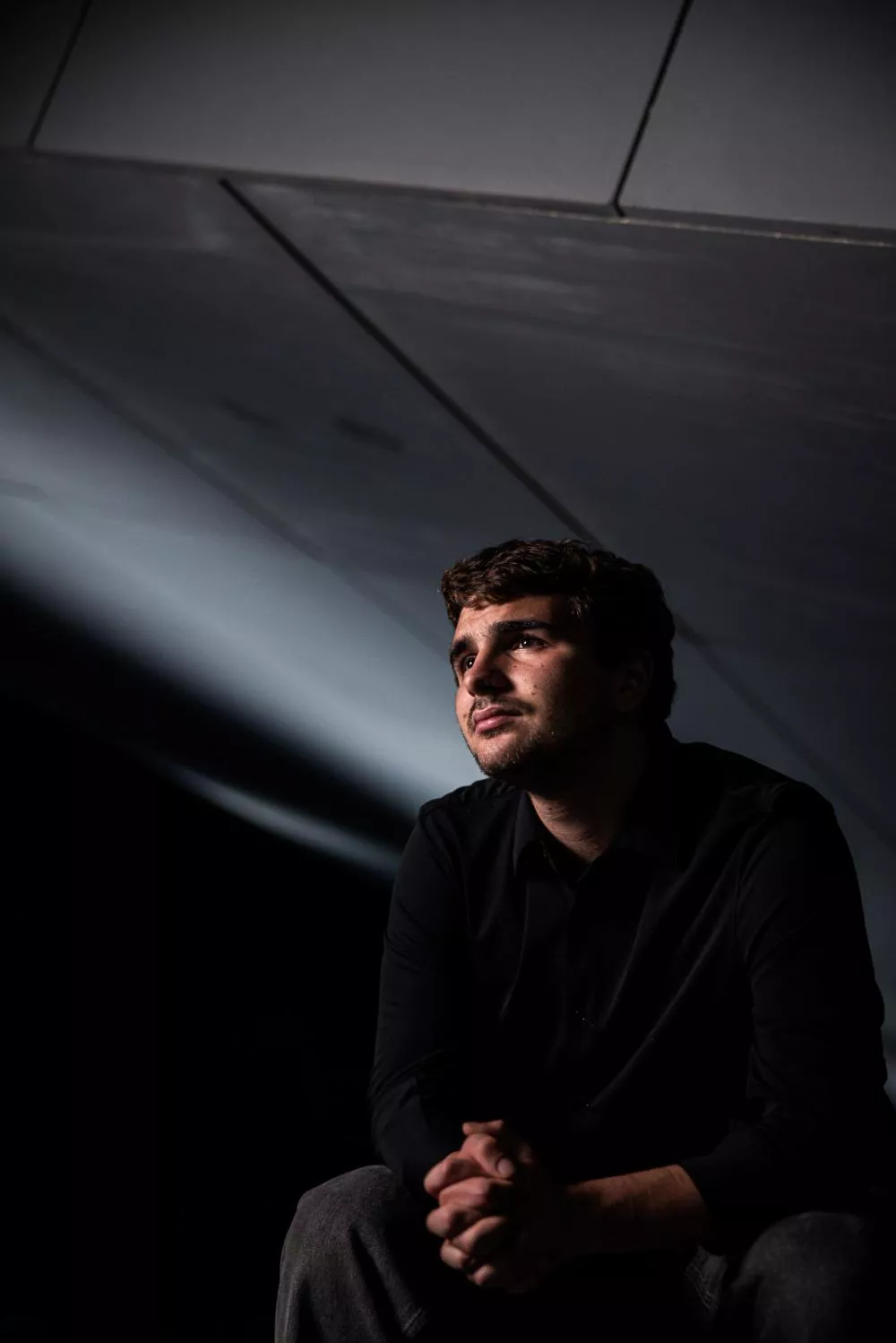 Un homme est assis, habillé en noir, regardant vers le haut, dans une ambiance sombre et éclairée de manière dramatique.
