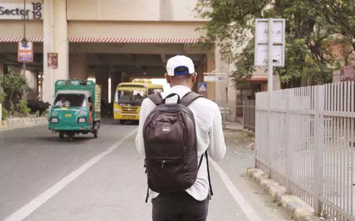 Un homme portant un sac à dos marche sur un trottoir à côté d'une route où circulent des véhicules, dont un tuk-tuk vert et un bus jaune. Il porte une chemise blanche et une casquette.
