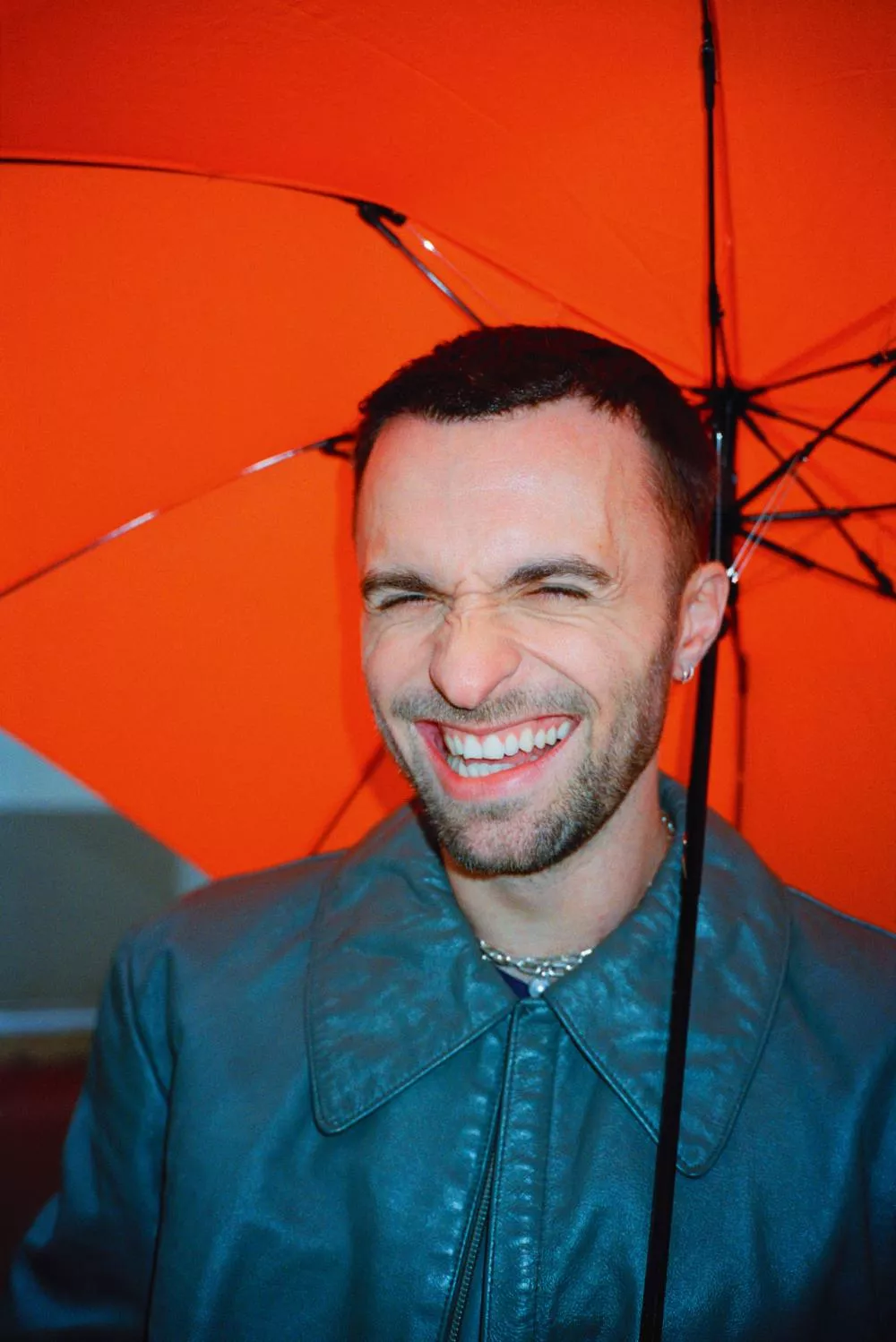 Un homme souriant se tient sous un parapluie orange, portant une veste en cuir bleu.