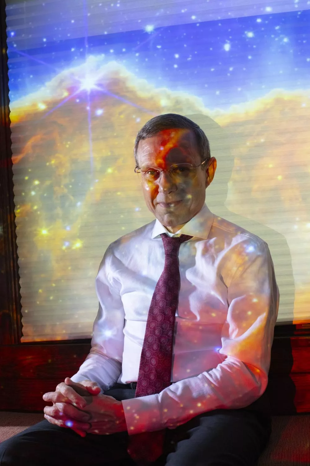 Un homme en chemise blanche et cravate rouge est assis, les mains croisées, avec une projection colorée de l'espace sur lui et en arrière-plan.