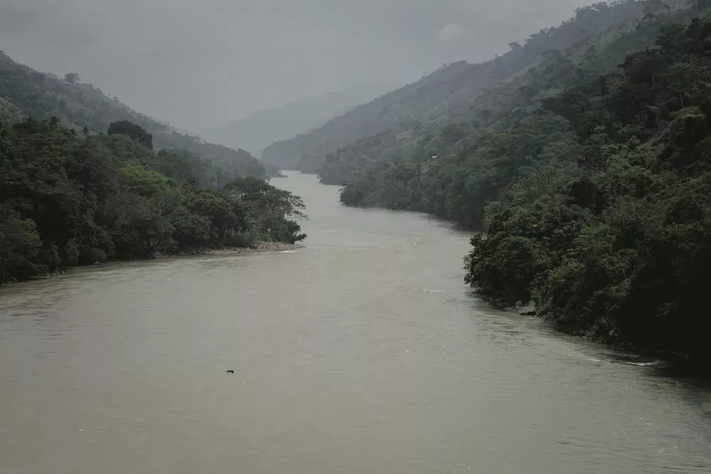 Un paysage naturel montrant une rivière sinueuse entourée de collines boisées sous un ciel nuageux.