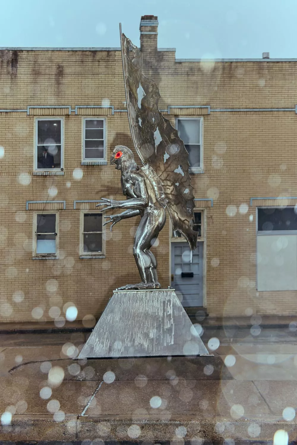 Une statue métallique représentant une créature humanoïde avec des ailes et des yeux rouges se tient sur un socle. Elle est située devant un bâtiment en briques jaunes.