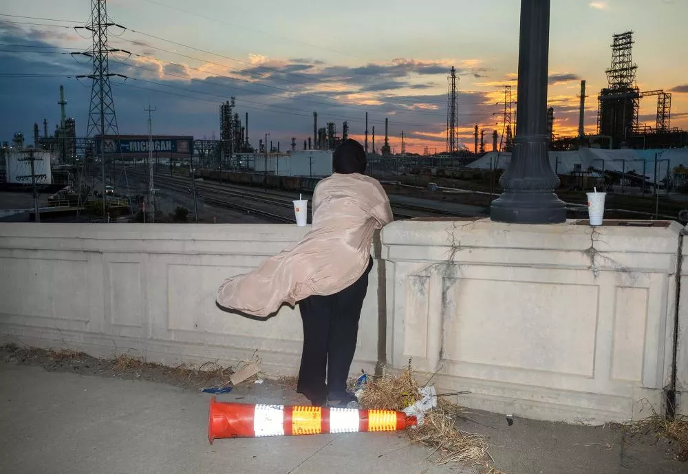 Une personne en manteau beige regarde un paysage industriel au coucher du soleil, appuyée sur un muret. Un cône de signalisation est renversé au sol, et un gobelet est posé sur le muret à côté d'un lampadaire.