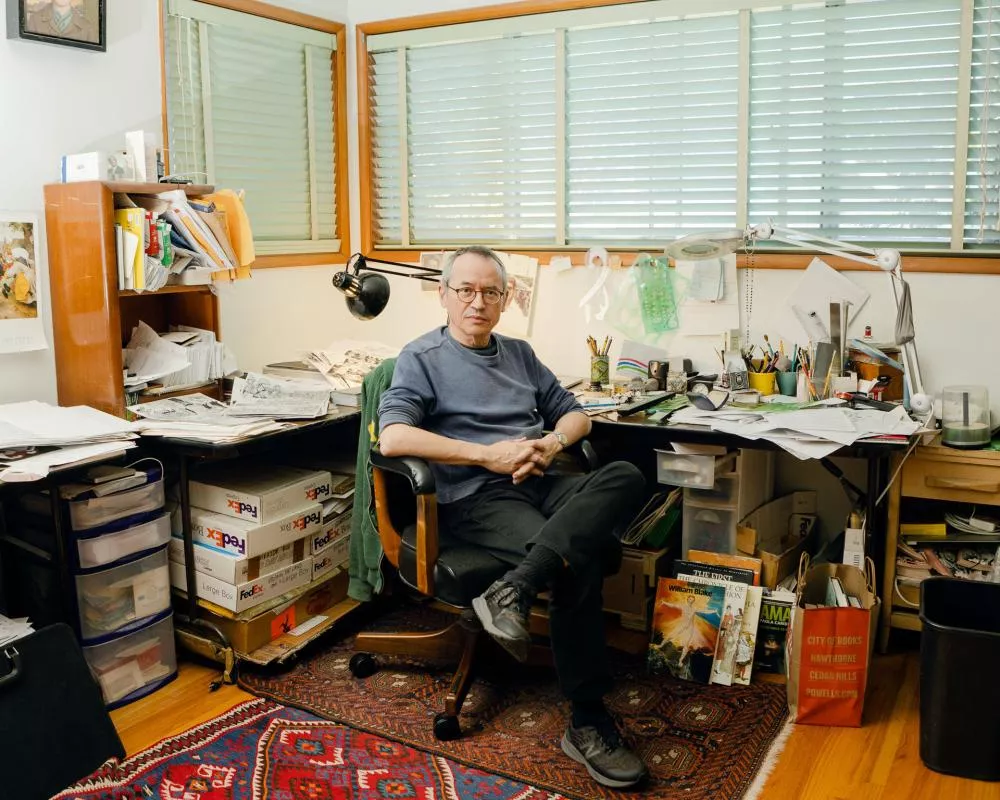 Un homme est assis dans un bureau encombré avec des papiers, des livres et des fournitures de bureau autour de lui. Il est entouré de fenêtres avec des stores et un tapis coloré recouvre le sol.