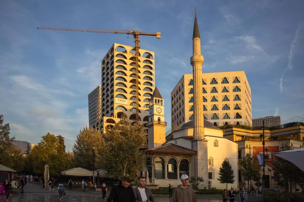 Une place urbaine avec des bâtiments modernes et historiques, dont une mosquée avec un minaret et une horloge. Des personnes se promènent dans la zone. Une grue de construction est visible à l'arrière-plan.