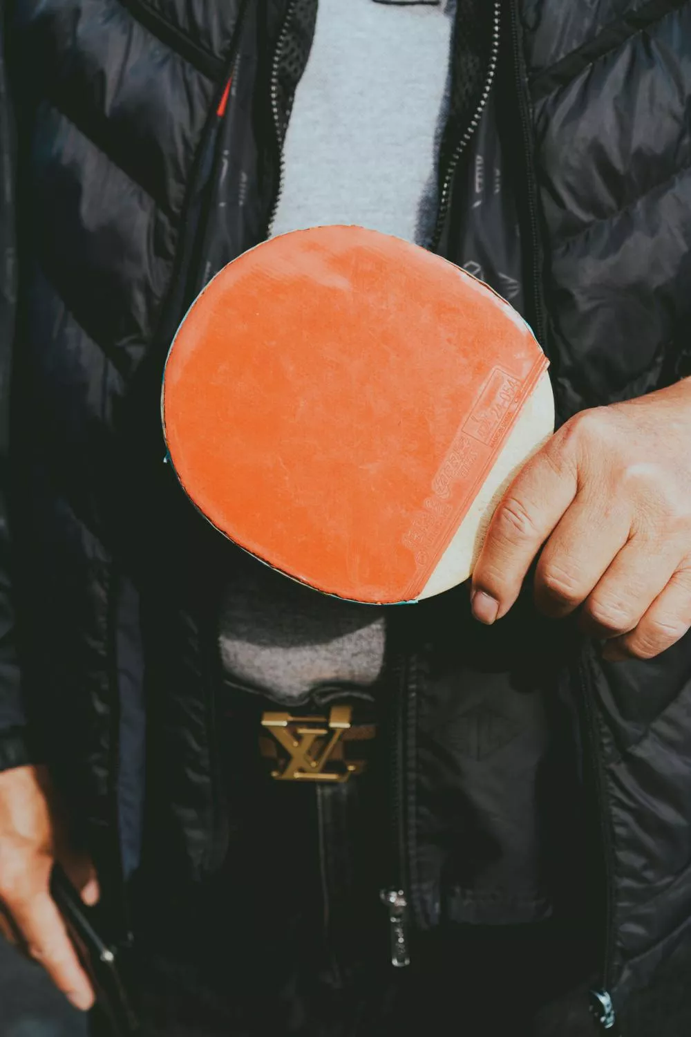 Un homme tient une raquette de ping-pong rouge. Il porte une veste noire et une ceinture avec une boucle dorée.