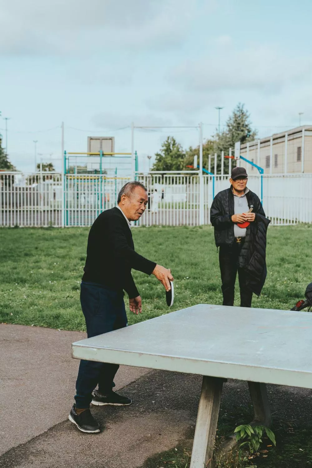 Deux personnes jouent au tennis de table en extérieur. L'une d'elles tient une raquette et semble en pleine action, tandis que l'autre observe en arrière-plan.