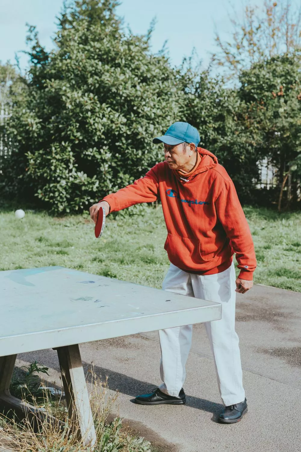 Un homme portant un sweat à capuche rouge et une casquette bleue joue au ping-pong en extérieur.