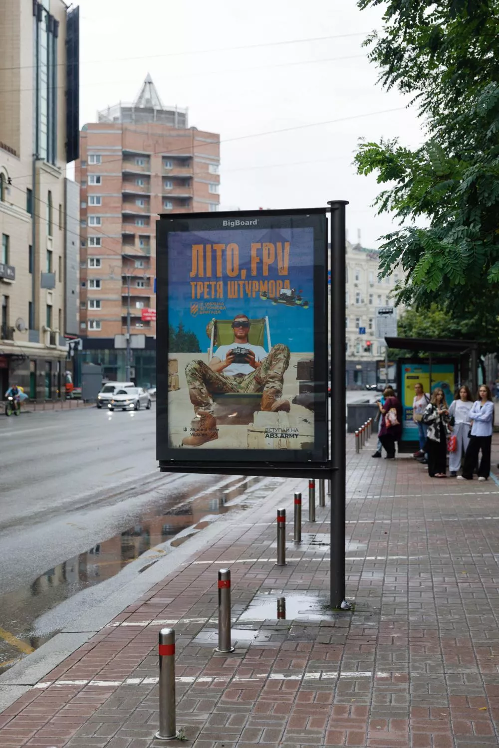 Une affiche publicitaire sur un panneau dans une rue montre un homme en tenue militaire assis, avec des inscriptions en ukrainien. Des passants attendent à un arrêt de bus à droite.