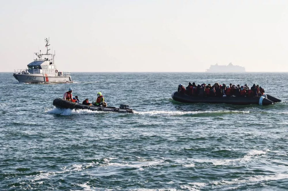 Des personnes portant des gilets de sauvetage se trouvent sur un canot pneumatique en mer, accompagnées par un bateau de sauvetage plus petit et un navire de patrouille à l'arrière-plan.
