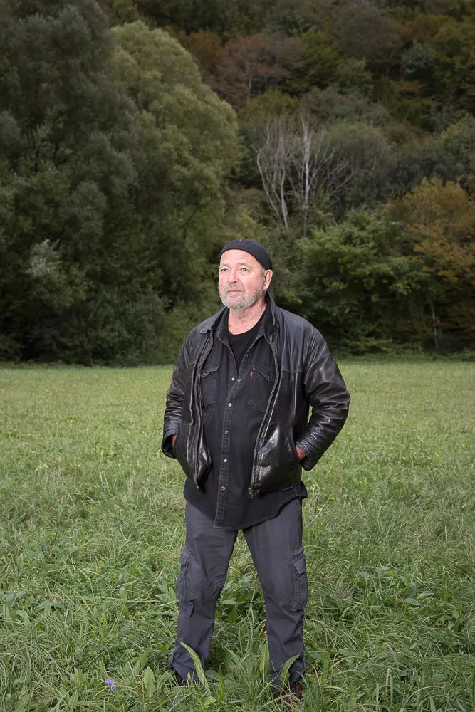 Un homme se tient debout dans un champ, portant une veste en cuir noire, une chemise sombre et un bonnet. L'arrière-plan montre des arbres et de la végétation.