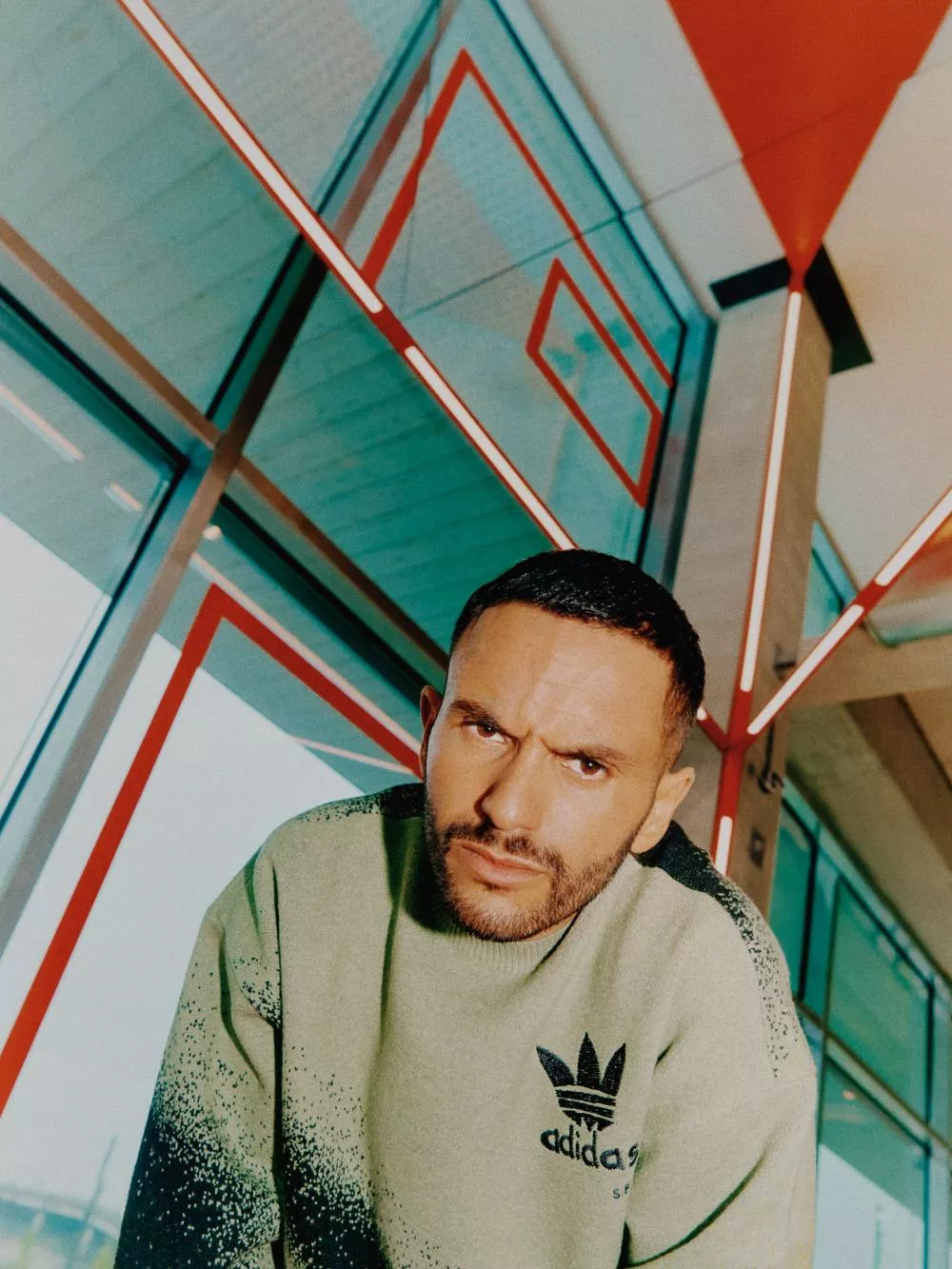 Un homme portant un pull vert Adidas se tient dans un environnement moderne avec des lignes rouges géométriques sur les murs et le plafond.