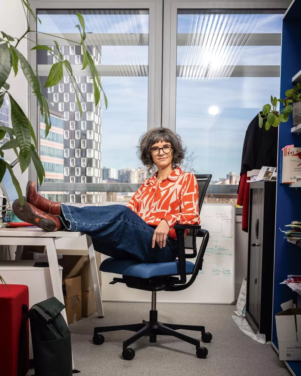 Une personne est assise dans un bureau, les pieds posés sur un bureau. Elle porte une chemise à motifs rouges et blancs, un jean et des bottes marron. Le bureau est entouré de plantes et de livres, avec une grande fenêtre donnant sur une vue urbaine en arrière-plan.