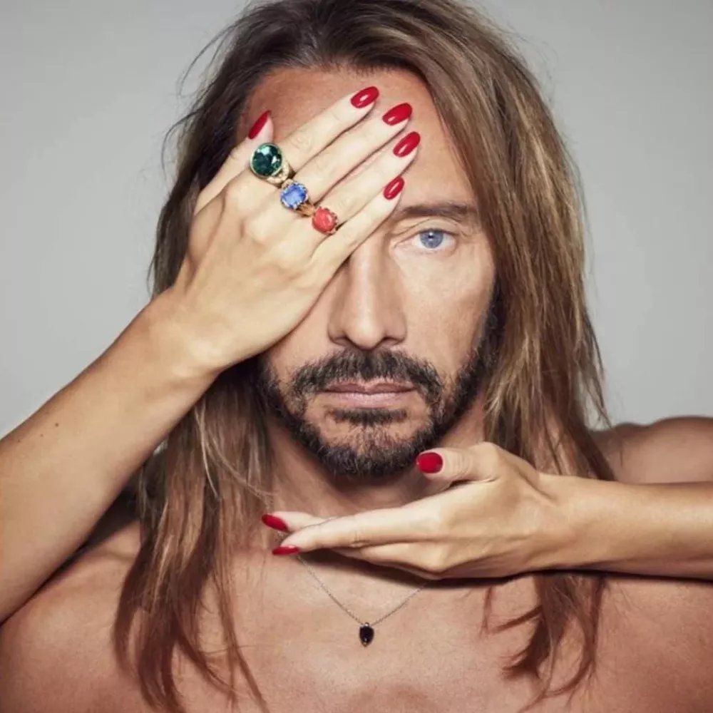 Un homme aux cheveux longs et à la barbe regarde droit devant. Une main féminine avec des ongles rouges et des bagues couvre un de ses yeux, tandis qu'une autre main est sous son menton.