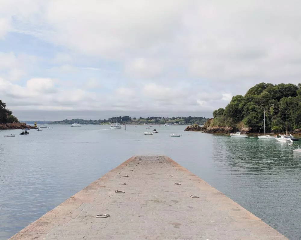 Un quai en pierre s'avance vers l'eau dans une baie. Des bateaux sont amarrés autour, avec des collines verdoyantes en arrière-plan sous un ciel partiellement nuageux.