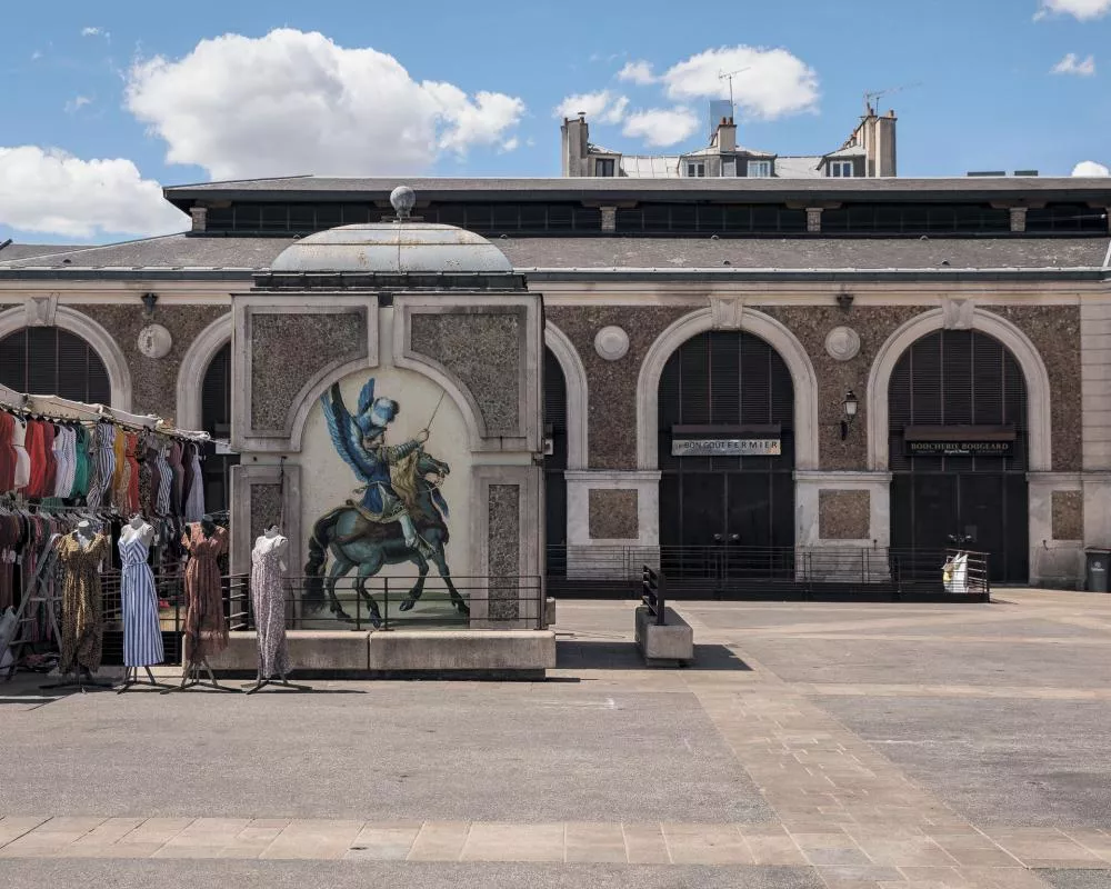Un bâtiment avec une façade en pierre et des arches, devant lequel se trouve une fresque représentant un personnage ailé sur un cheval. À gauche, plusieurs mannequins exposent des vêtements colorés. Le ciel est bleu avec des nuages.