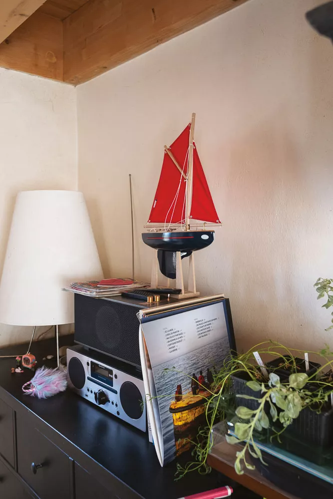 Une table avec une lampe, une maquette de bateau à voile rouge, des livres, une radio, et une plante en pot.