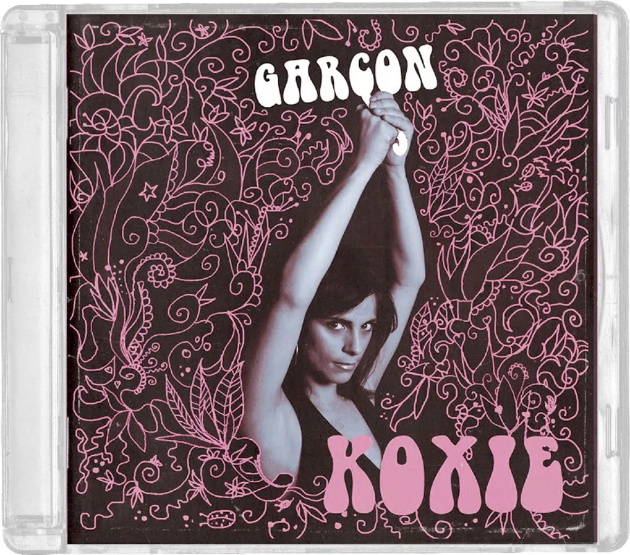 La couverture d'un album avec un fond noir orné de motifs floraux roses. Une femme avec les bras levés est au centre, et les mots "GARÇON" et "KOXIE" apparaissent en lettres blanches et roses.