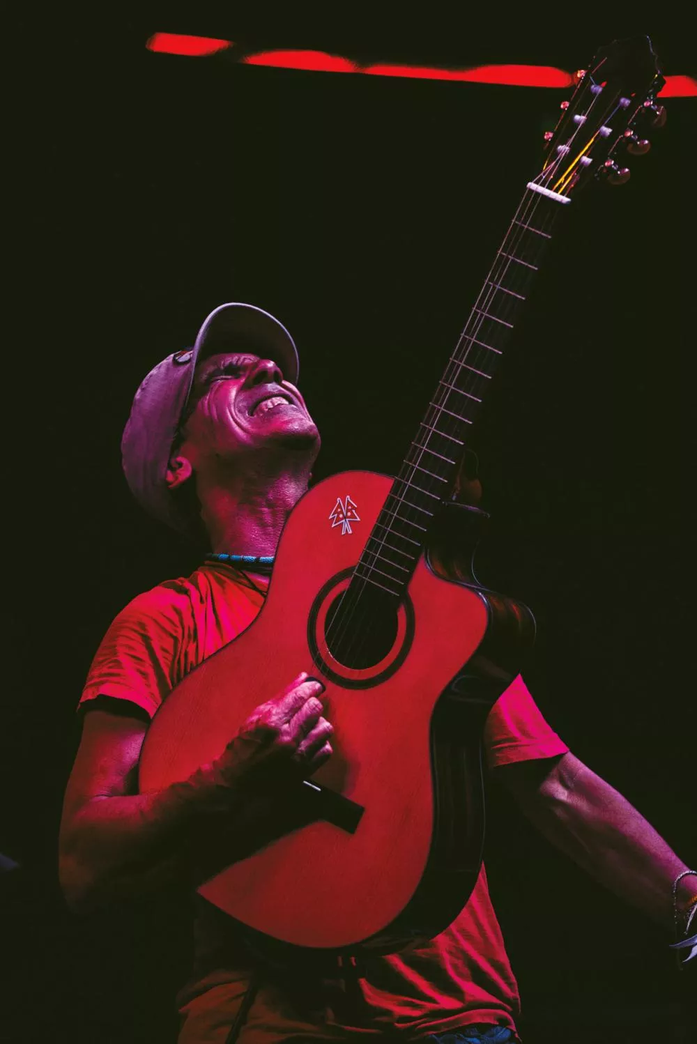 Un homme portant une casquette joue de la guitare avec passion, affichant un sourire enjoué. La scène est éclairée par une lumière rouge, créant une ambiance intense.