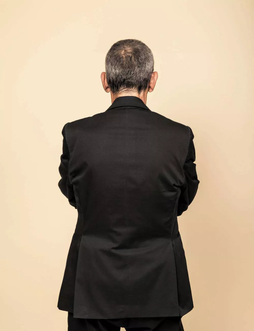 Un homme en costume noir vu de dos, sur un fond beige.