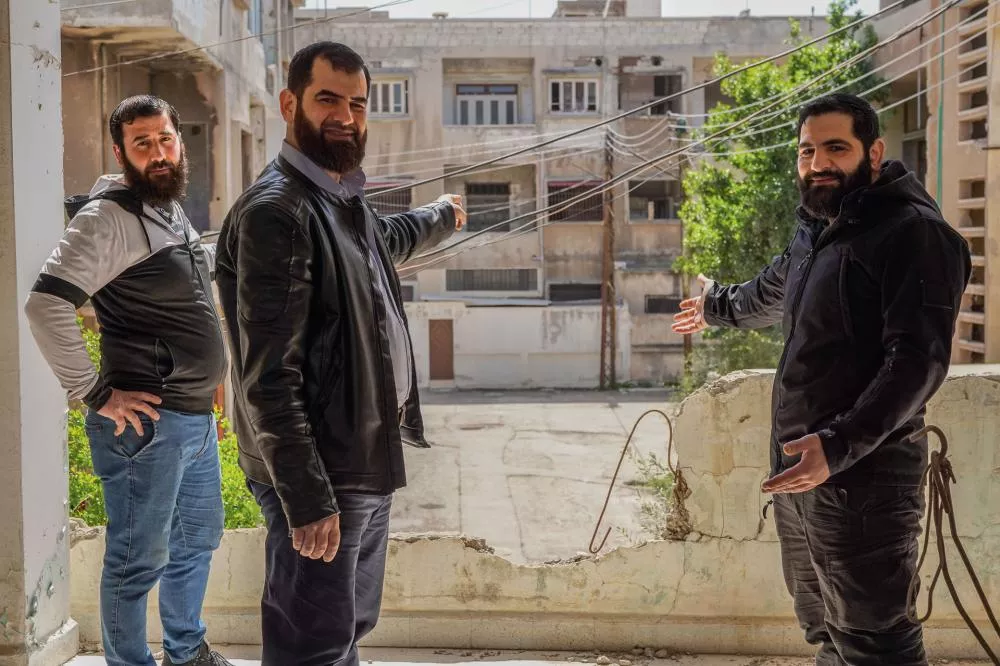 Trois hommes barbus se tiennent sur un balcon délabré, pointant vers un bâtiment en face. Ils sont habillés de manière décontractée et sourient légèrement. L'arrière-plan montre un environnement urbain avec des bâtiments usés et des câbles électriques visibles.
